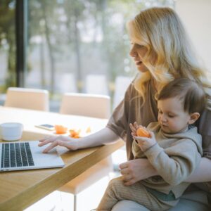 A mother works on a laptop at home while holding her baby, showcasing remote working and parenting.
