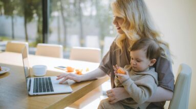 A mother works on a laptop at home while holding her baby, showcasing remote working and parenting.